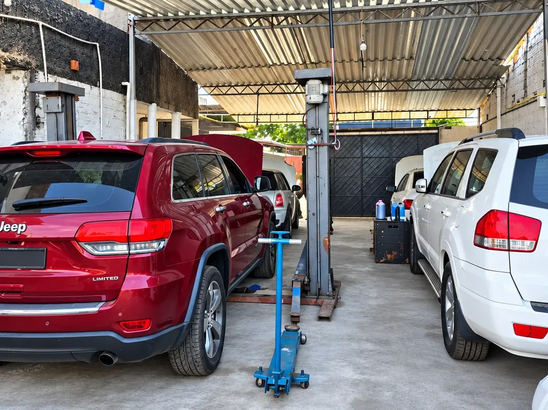Área coberta da oficina com o elevador, um macaco hidráulico e carros em atendimento, entre eles um Jeep Grand Cherokee vermelho.